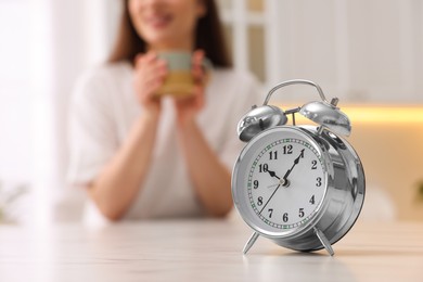 Photo of Alarm clock on white table. Woman with cup of drink in kitchen, selective focus