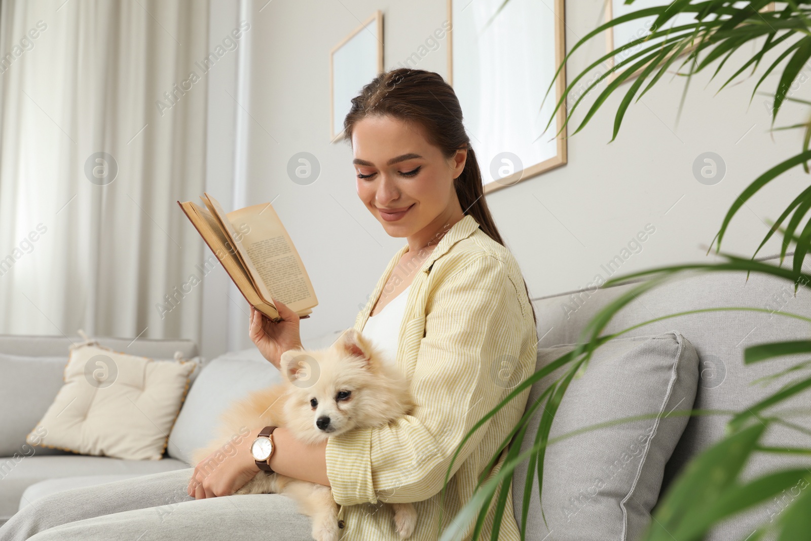 Photo of Happy young woman reading book with cute dog on sofa in living room