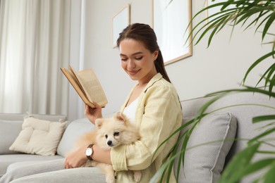 Happy young woman reading book with cute dog on sofa in living room