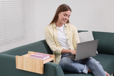 Happy woman using laptop on sofa with wooden armrest table at home