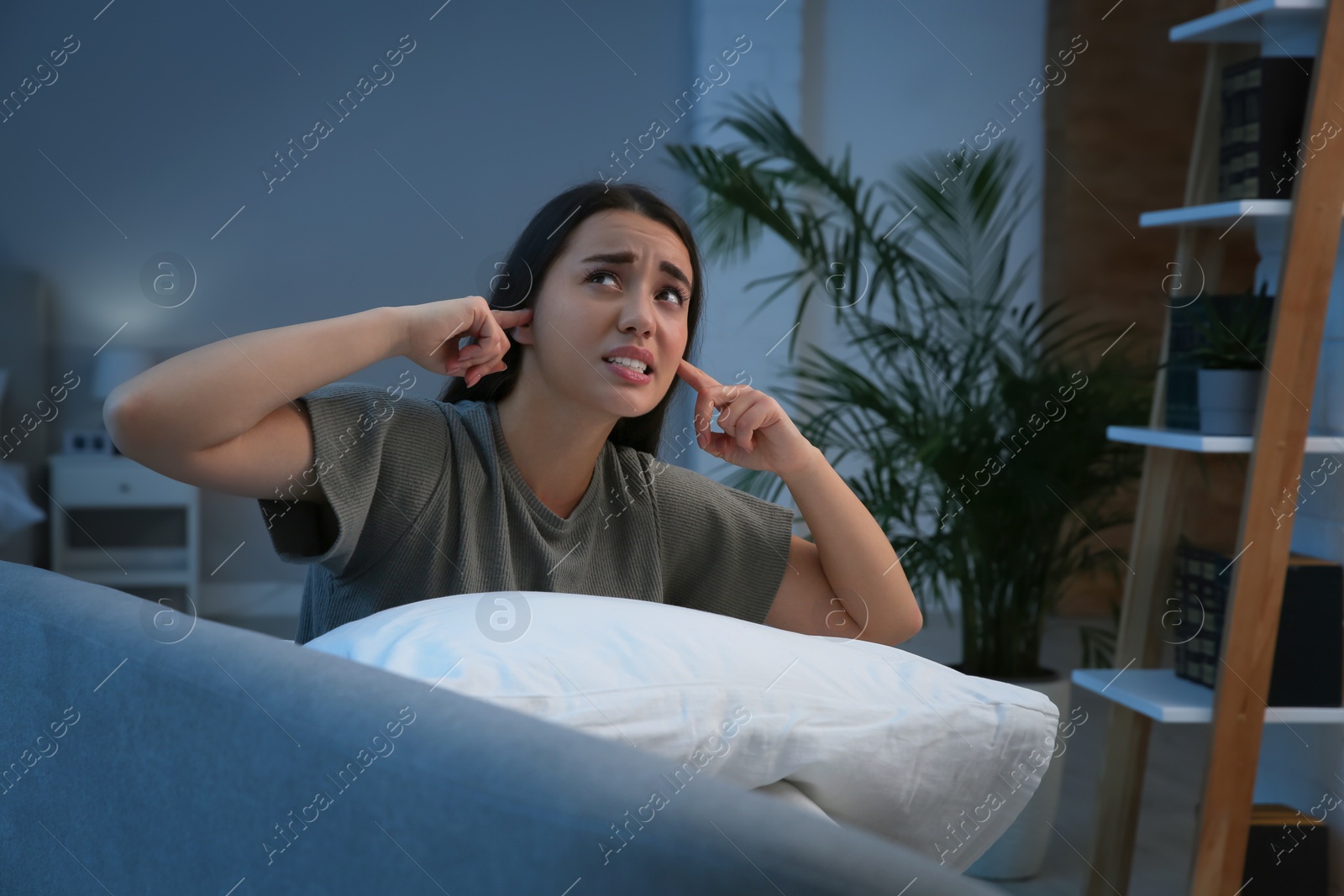 Photo of Unhappy young woman covering ears in living room at night. Noisy neighbours