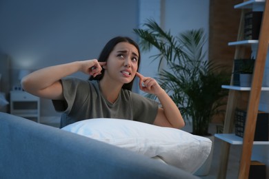 Photo of Unhappy young woman covering ears in living room at night. Noisy neighbours