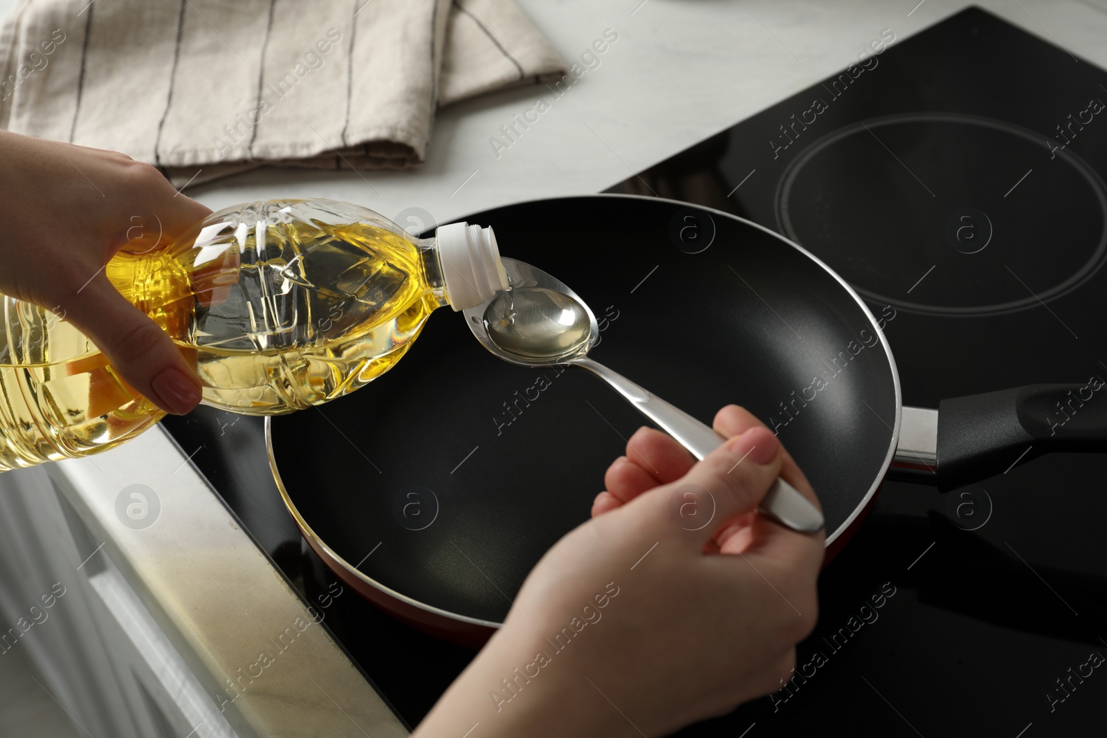 Photo of Woman with spoon pouring cooking oil into frying pan on stove, closeup