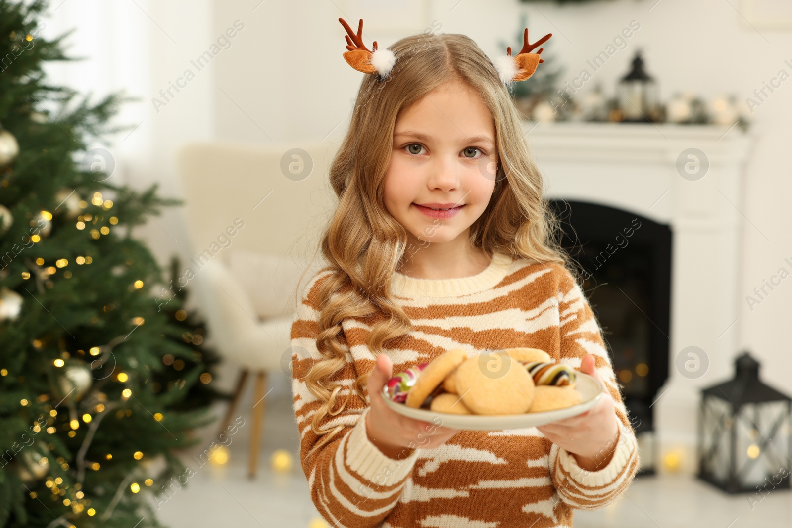 Photo of Portrait of cute little girl in Christmas hair clips holding plate with cookies at home