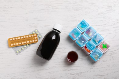 Photo of Bottle of syrup, measuring cup, weekly pill organizer and pills on white wooden table, flat lay. Cold medicine