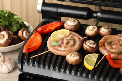 Photo of Electric grill with homemade sausages, mushrooms and bell pepper on table, closeup