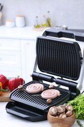 Photo of Electric grill with homemade sausages, mushrooms, and vegetables on white table in kitchen