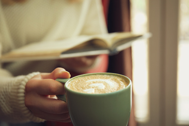 Woman with coffee reading book indoors, focus on cup