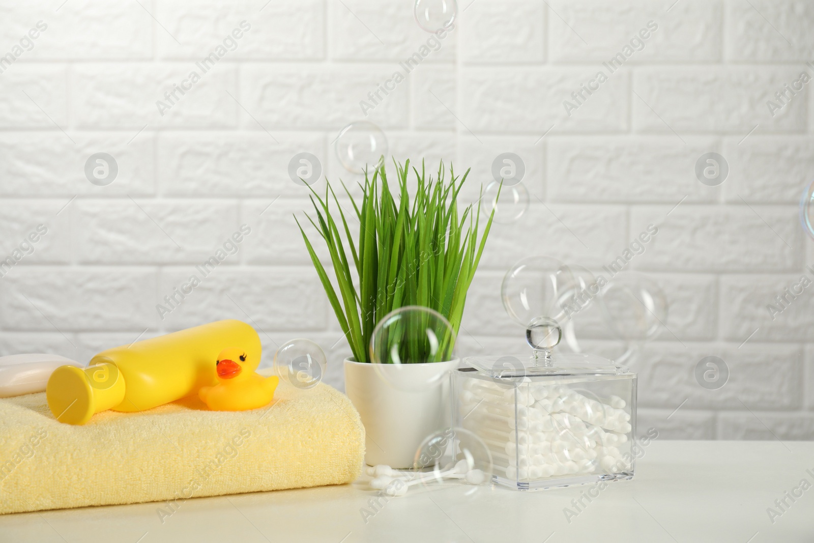 Photo of Baby bath accessories. Towel, cosmetic products, cotton swabs and toy duck on white table against brick wall