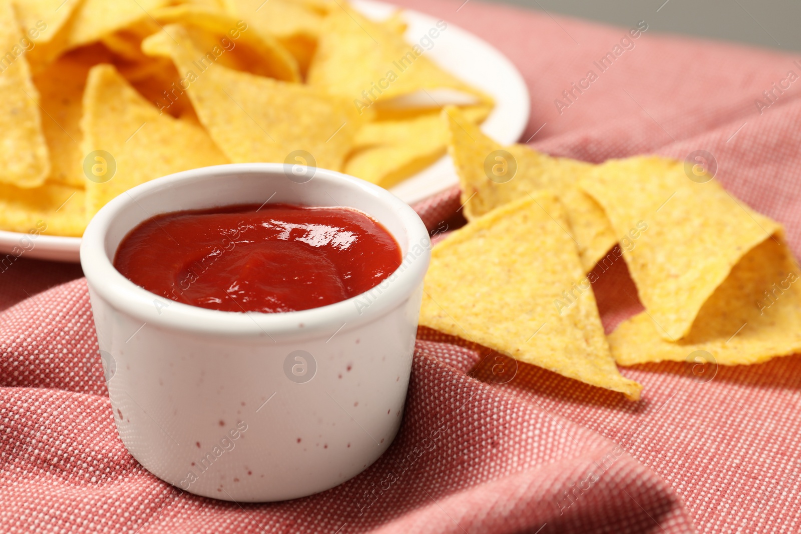 Photo of Tasty ketchup and tortilla chips on red tablecloth, selective focus