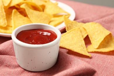 Photo of Tasty ketchup and tortilla chips on red tablecloth, selective focus