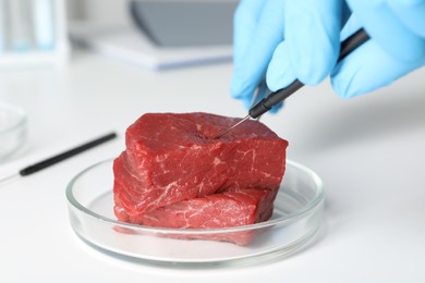Scientist with dissecting needle examining piece of raw cultured meat at white table, closeup