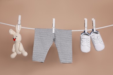 Photo of Cute small baby shoes, pants and toy hanging on washing line against brown background