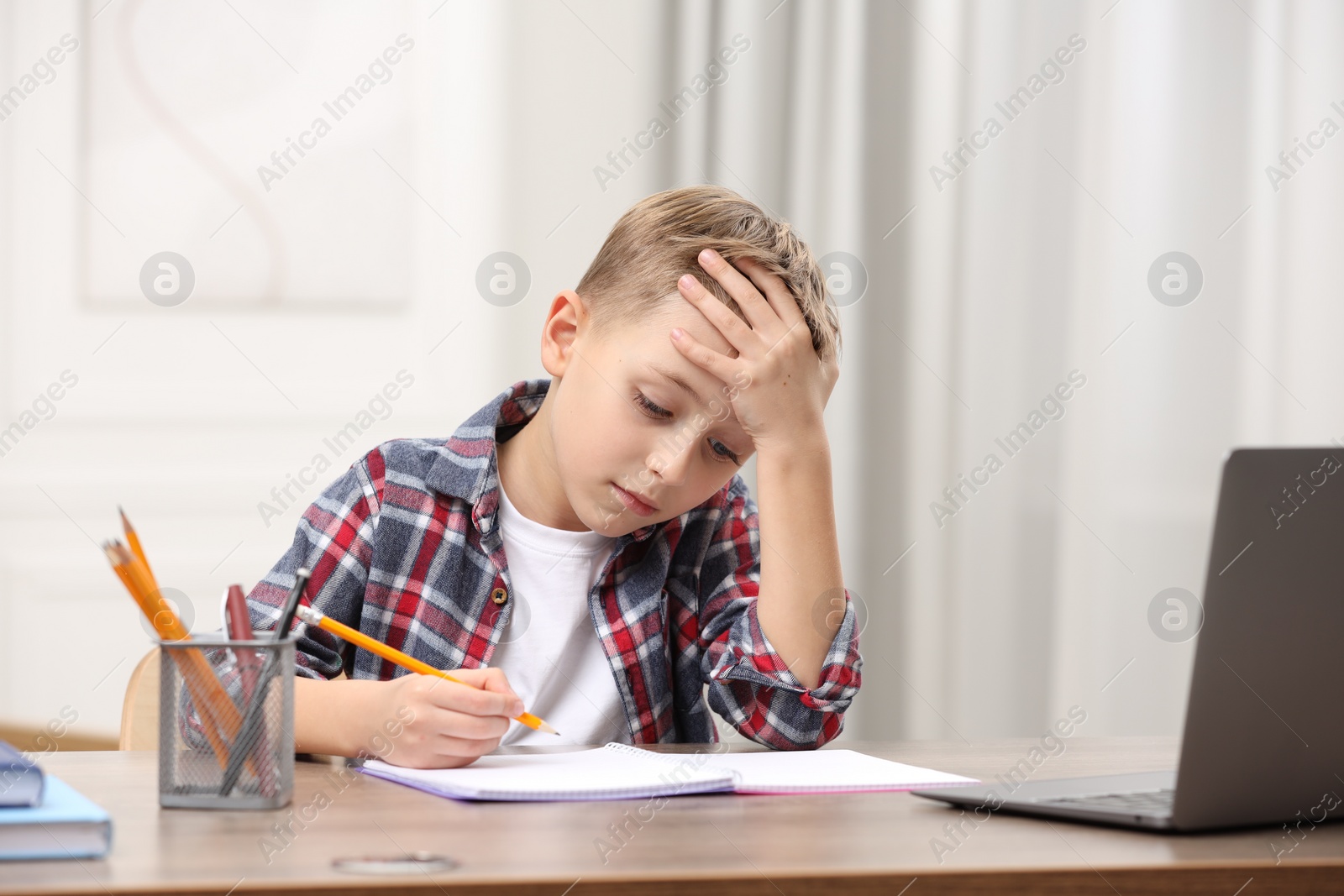 Photo of Little boy suffering from headache while studying at wooden desk indoors