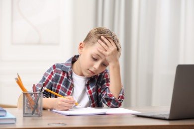 Photo of Little boy suffering from headache while studying at wooden desk indoors