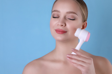 Young woman washing face with cleansing brush on light blue background. Cosmetic product