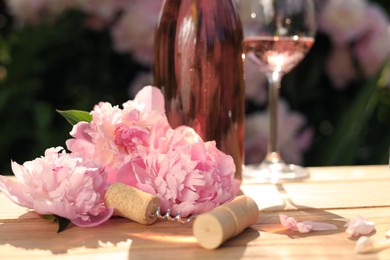 Rose wine and corkscrew near beautiful peonies on wooden table in garden, closeup