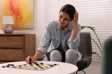 Upset woman playing checkers in armchair at home