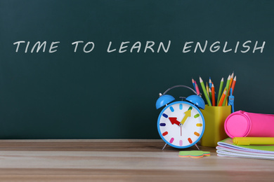 Alarm clock and stationery on table near green chalkboard with text Time to Learn English