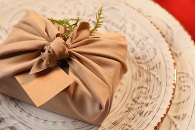 Photo of Furoshiki technique. Gift packed in beige fabric with blank card on plate, closeup