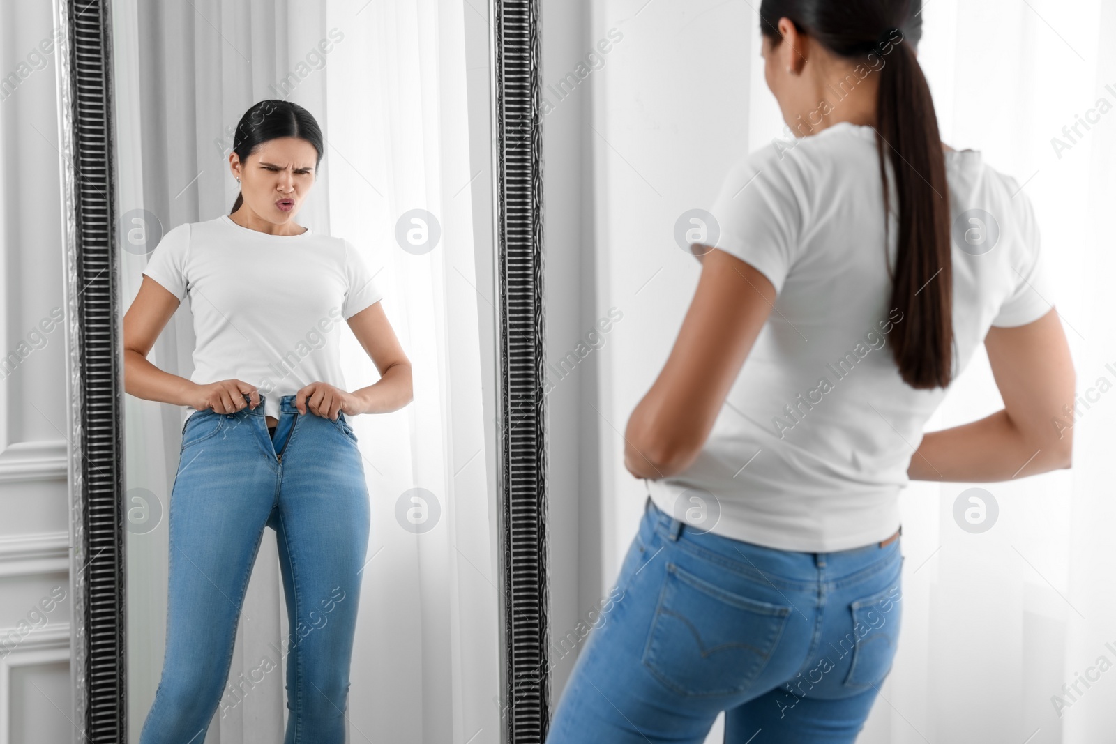 Photo of Young woman trying to put on tight jeans near mirror indoors