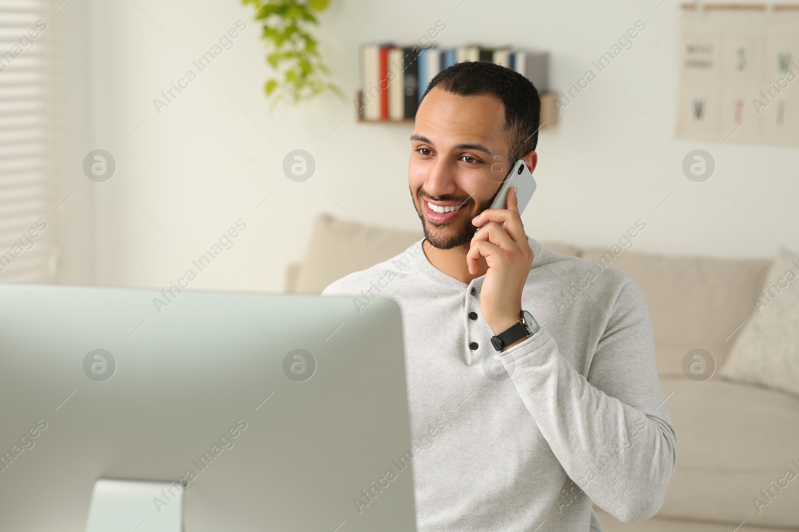 Photo of Young man talking on smartphone while working with computer in room. Home office