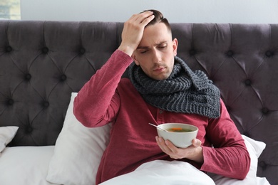 Sick young man eating broth to cure cold in bed at home