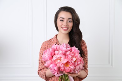 Beautiful young woman with bouquet of pink peonies near white wall