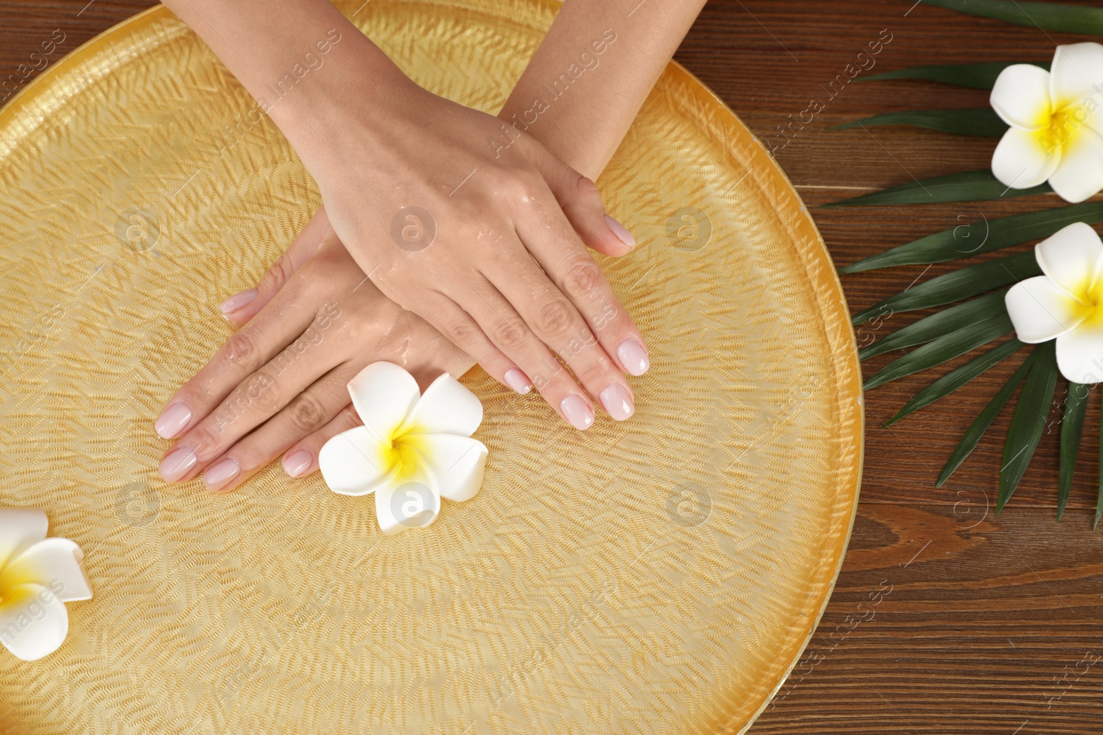 Photo of Woman soaking her hands in bowl with water and flowers on wooden table, top view. Spa treatment