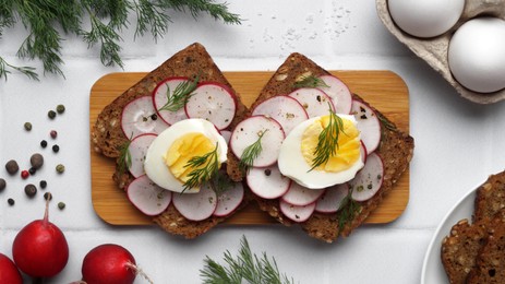 Tasty sandwiches with boiled egg, radish and ingredients on white tiled table, flat lay