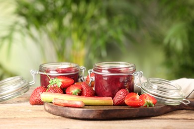 Tasty rhubarb jam in jars, stems and strawberries on wooden table against blurred background. Space for text