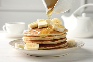 Photo of Pouring honey from jug onto delicious pancakes with bananas and butter at white wooden table, closeup