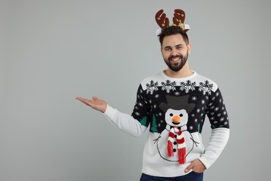Photo of Young man in Christmas sweater and reindeer headband showing something on grey background. Space for text