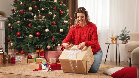 Photo of Beautiful young woman wrapping gift near Christmas tree at home