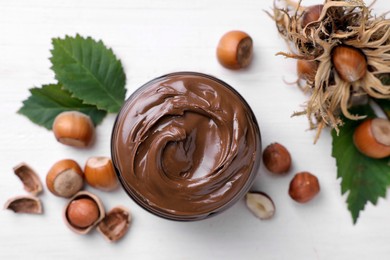 Photo of Jar with tasty chocolate spread, hazelnuts and green leaves on white wooden table, flat lay