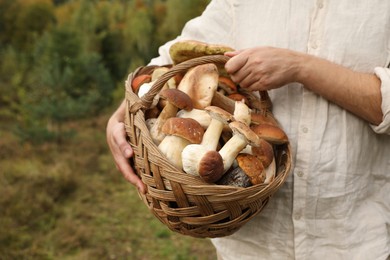 Photo of Man holding wicker basket with fresh wild mushrooms outdoors