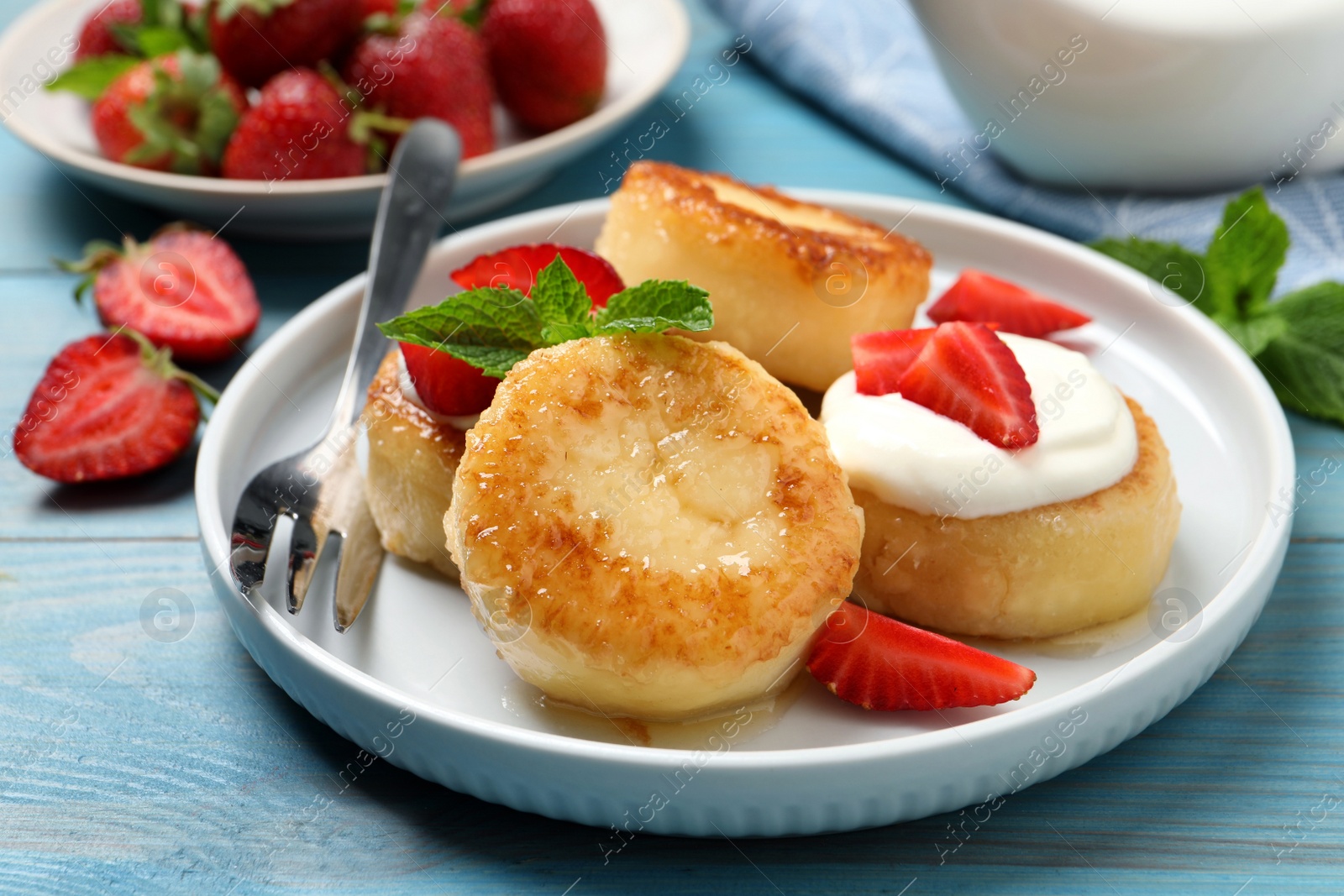 Photo of Delicious cottage cheese pancakes with strawberries, honey and sour cream on light blue wooden table, closeup