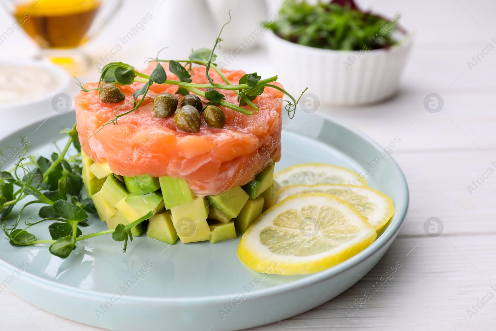 Photo of Tasty salmon tartare with avocado, lemon, capers and microgreens on white wooden table, closeup