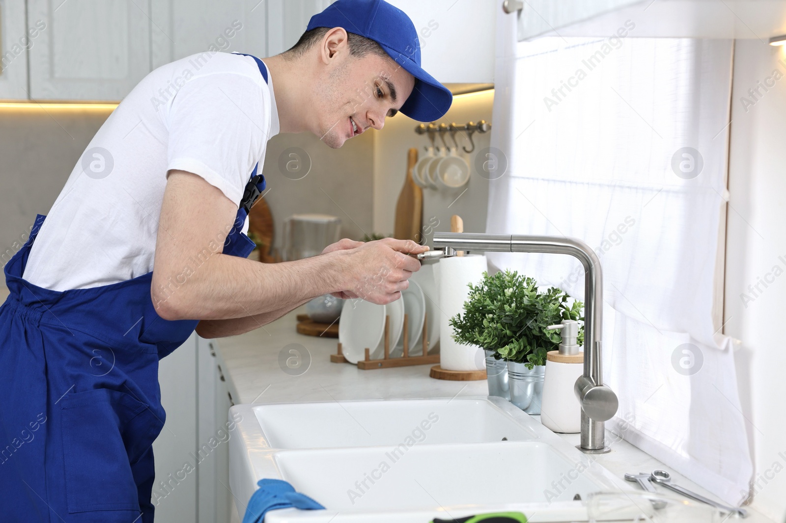 Photo of Smiling plumber repairing faucet with spanner in kitchen