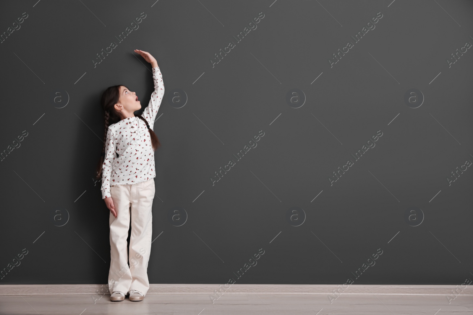 Photo of Emotional little child measuring her height near black wall indoors, space for text
