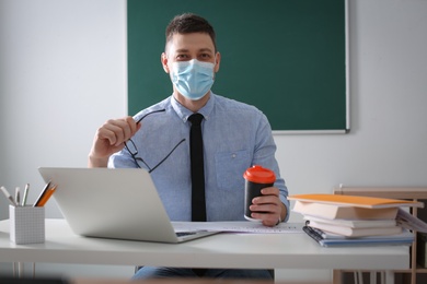Photo of Teacher with protective mask and laptop sitting at desk in classroom. Reopening after Covid-19 quarantine