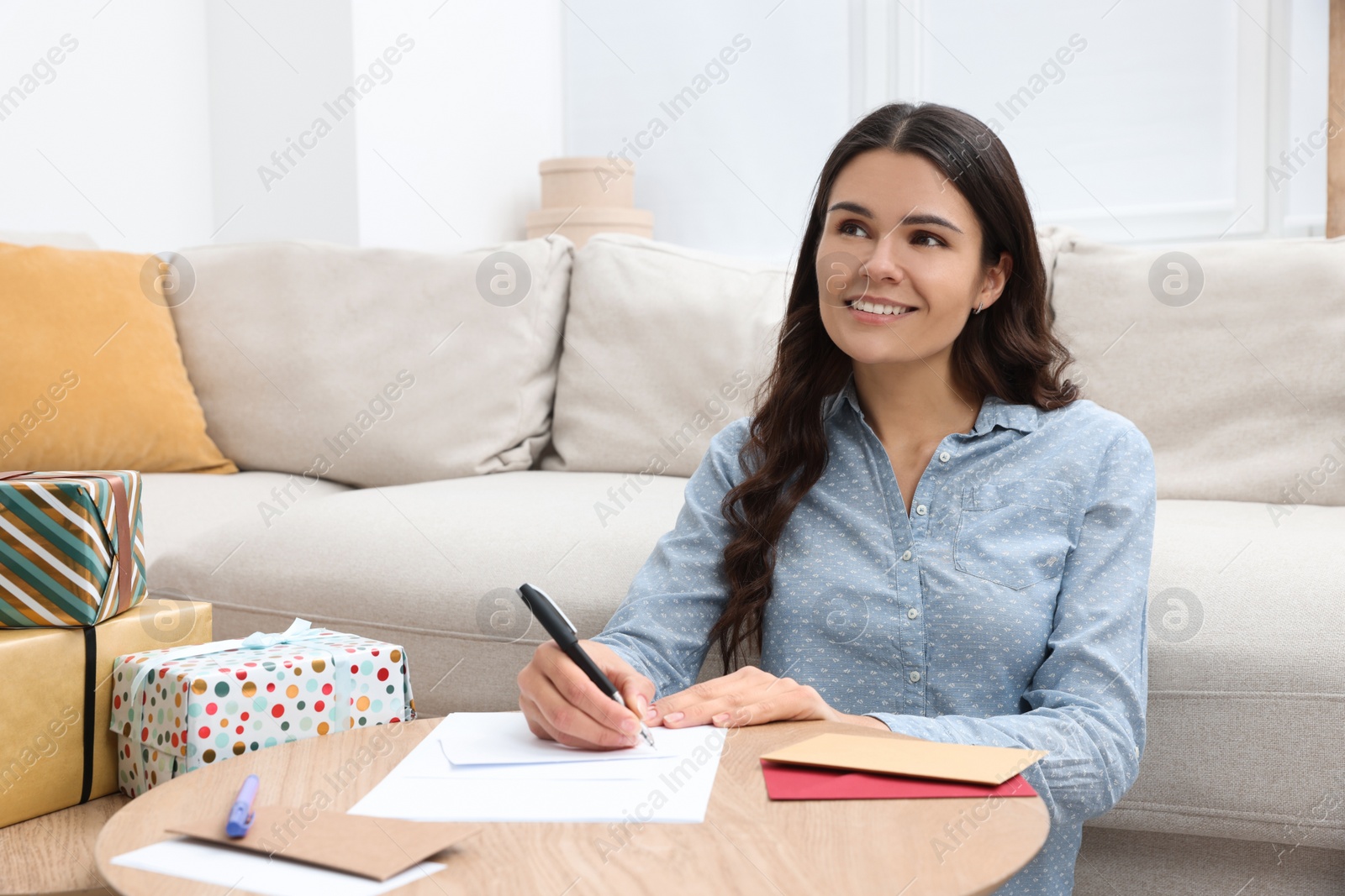 Photo of Young woman writing message in greeting card at table indoors