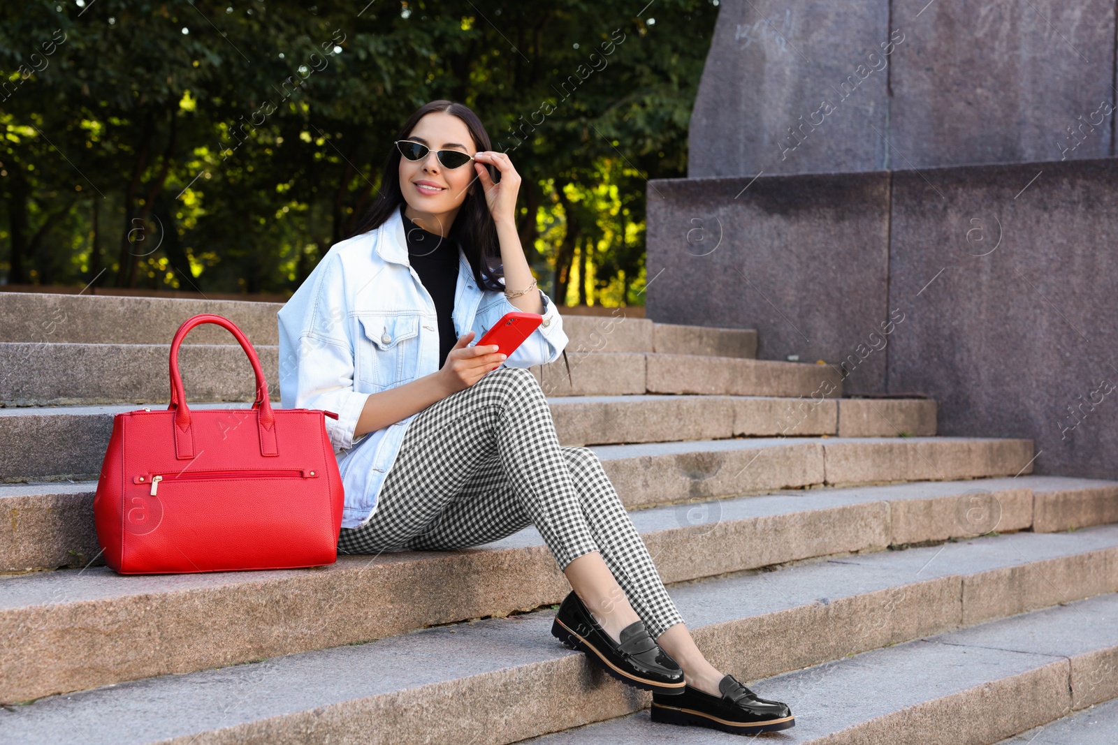 Photo of Young woman with stylish bag and smartphone sitting on stairs outdoors, space for text