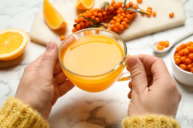 Woman with cup of fresh sea buckthorn tea at white marble table, closeup