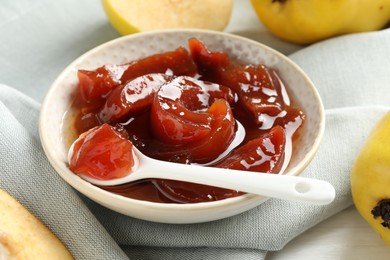 Photo of Tasty homemade quince jam in bowl, spoon and fruits on table, closeup