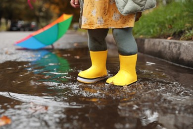 Photo of Little girl splashing water with her boots in puddle outdoors, closeup