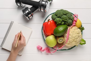 Photo of Woman developing diet plan at white wooden table with products and dumbbells, top view