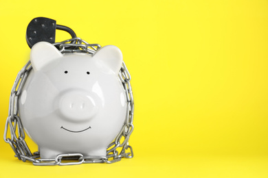 Photo of Piggy bank with steel chain and padlock on yellow background, space for text. Money safety concept