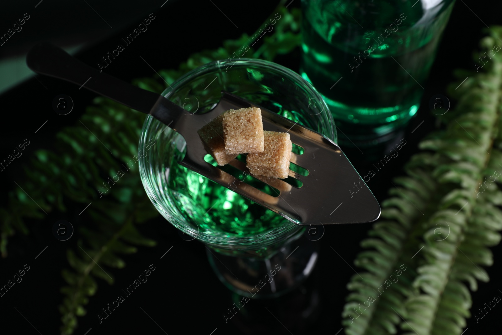 Photo of Absinthe in glass, brown sugar, spoon and green leaves on mirror table, top view. Alcoholic drink