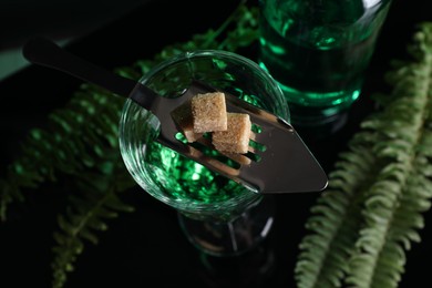 Photo of Absinthe in glass, brown sugar, spoon and green leaves on mirror table, top view. Alcoholic drink